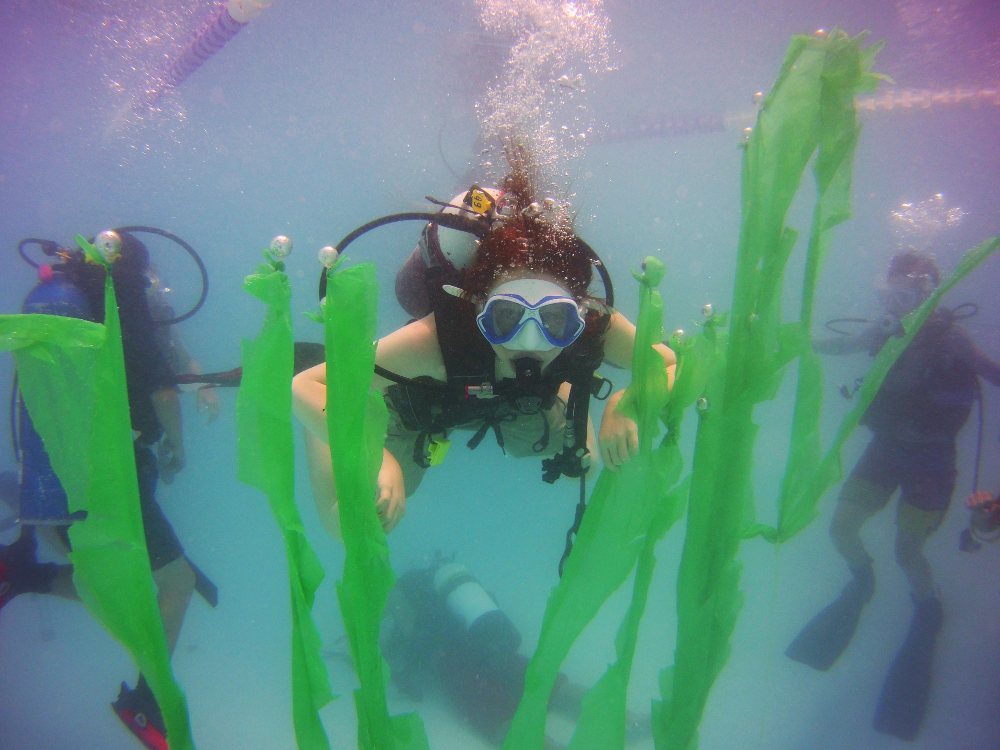 A young diver looks at the camera while wearing scuba diving gear and swimming through imitation kelp.