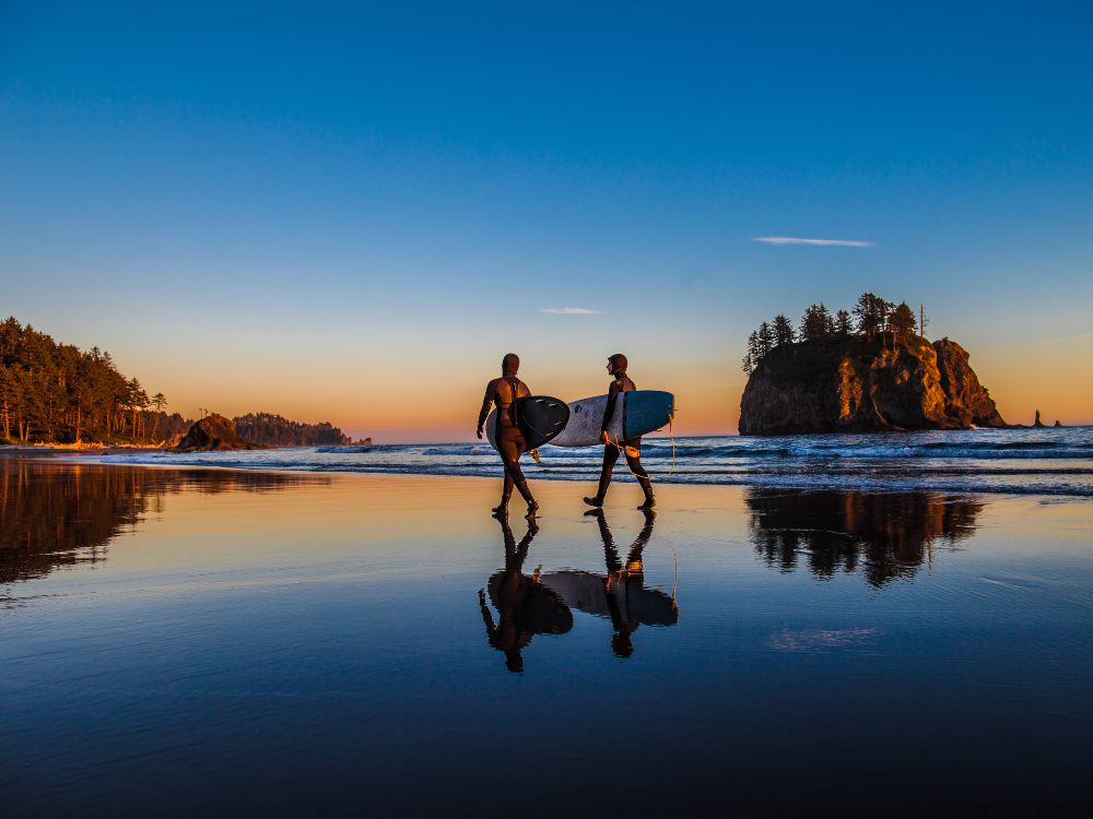 Surfers walk along a beach at sunset in Olympic Coast National Marine Sanctuary.