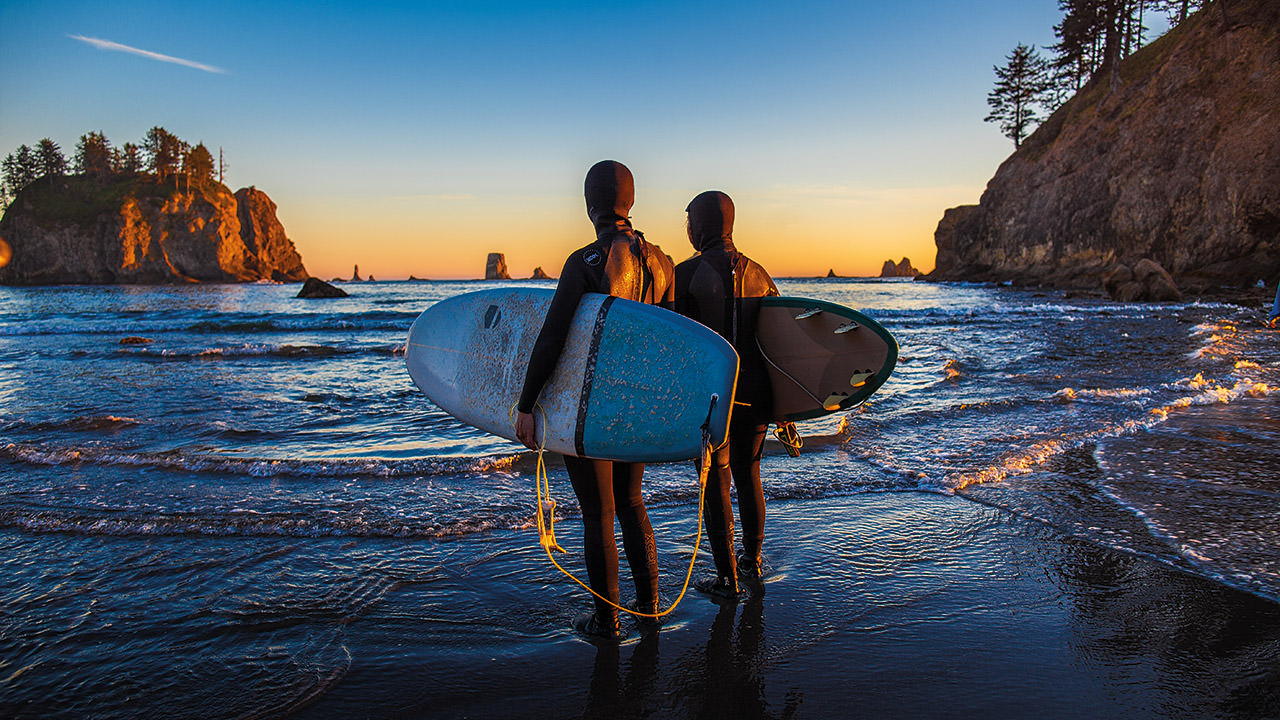 a surfer and bodyboarder standing on the beach looking at the water