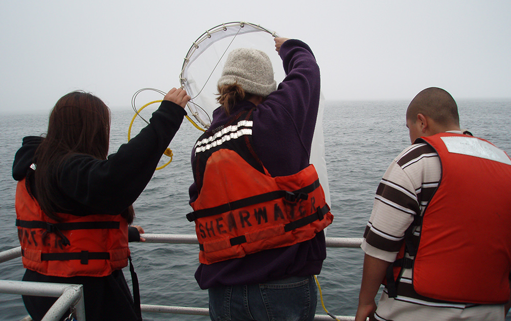 three students on a vessel face toward the ocean and pull up a net
