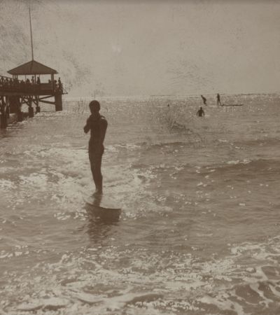 A stereograph postcard shows a man surfing in Honolulu Harbor in 1915.