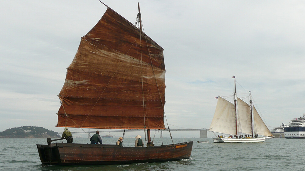 a wooden vessel at full sail with another sailboat visible in background