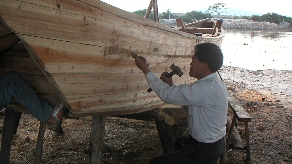 a person sitting on a work bench in front of a wooden boat that is lifted off the ground by support beams. The person is holding a hammer in one hand