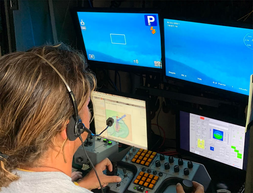 a person staring at two computer screens that show imagery of an underwater shipwreck. The person is holding the controls that move the remotely operated vehicle around