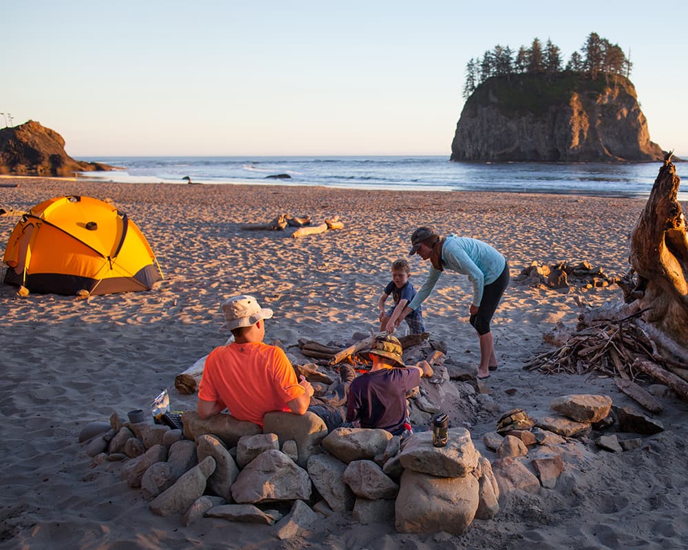 people gather next to a fire pit on the beach. There is a tent in the foreground and the ocean in the background.