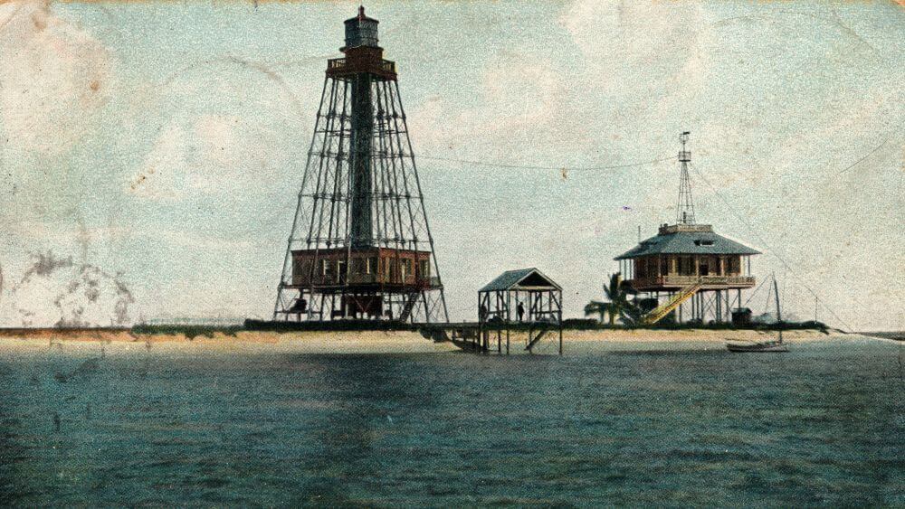 a lighthouse with an open metal frame and a lightkeeper's house on stilts sit on a low sandy island