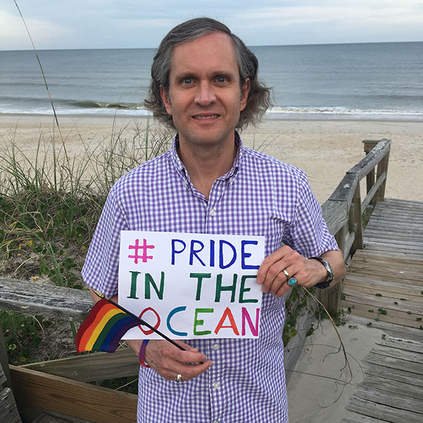 A person standing in front of the ocean holding a pride flag with a sign that reads '# Pride In The Ocean.'