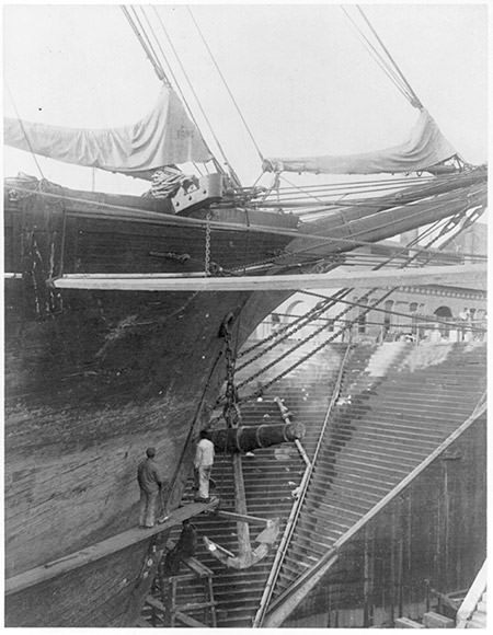 ship builders standing on a plank attach the hull of a ship, a cannon is being raised